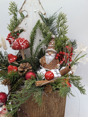 Weihnachtsmann-Figur mit brauner Mütze zwischen Tannengrün, roten Pilzen, Beeren, Zimtstangen und Päckchen