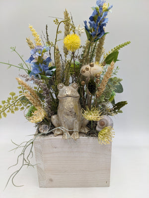 Sommergesteck in heller Holzlade mit goldenem Frosch, blauen Blüten, gelben Blütenköpfen und Ähren