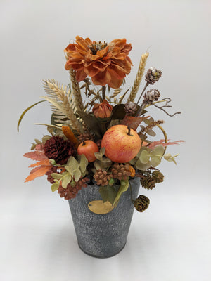 Herbstgesteck im grauen Zinktopf mit oranger Blüte, Äpfeln, Weizenähren, Zapfen und Herbstblättern