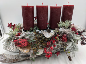 Adventsgesteck mit vier bordeauxroten Stumpenkerzen, roten Kugeln, Beeren und Zapfen auf beschneitem Holzschlitten