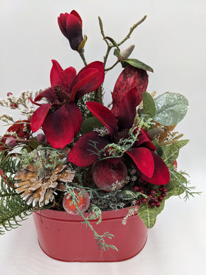 Nahaufnahme des Wintergestecks: rote Magnolienblüten, Beeren, glitzerndes Blatt und getrocknete Banksia über roter Schale