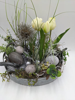 Ostergesteck von vorn: weiße Tulpen, dunkles Glitzerei, gesprenkeltes Ei, Beeren und Blattzweige in Metallschale