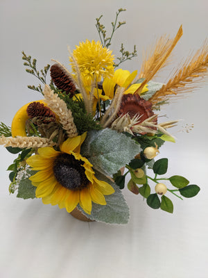 Herbstgesteck von der Seite: zwei gelbe Sonnenblumen, rostbraune Blüte, gelber Kürbis, Weizen und Gräser