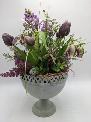 Frühlingsgesteck von vorn: lila Hyazinthen, Tulpen, Schachbrettblumen, Wachtelei und Bast im grauen Pokal
