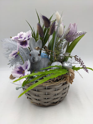 Ostergesteck im grauen Weidenkorb mit grauem Hasen, lila Tulpen, weiß-violetten Stiefmütterchen und Federn