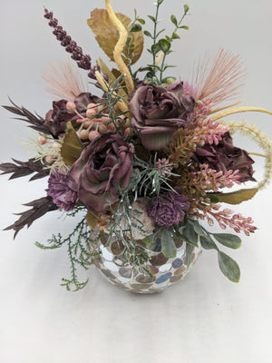 Gesteck von oben: lila Rosen, rosa Heidekraut, helle Gräser und dunkelrote Blätter über der Mosaikvase