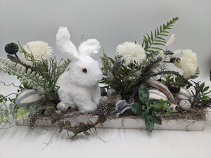 Längliches Ostergesteck mit weißem Hasen, Craspedia, Farn und grau gestreiften Eiern in weißem Holztablett