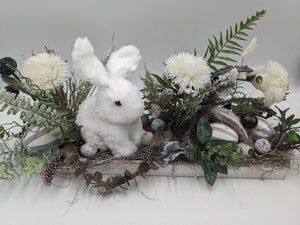 Ostergesteck von vorn: weißer Hase zwischen cremeweißen Blüten, Federn, Nest und gesprenkelten Eiern