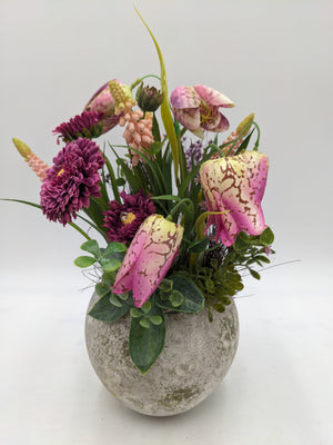 Gesteck mit rosa Schachbrettblumen, lila Bellis und Gräsern in rundem Topf in Steinoptik