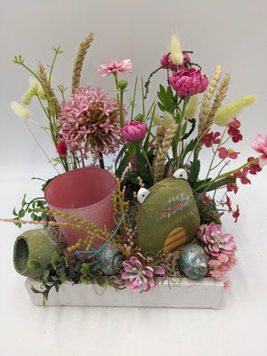 Nahansicht des Gestecks: Froschfigur mit gestreiftem Ei, rosa Sukkulenten, blaugrüne Schnecken und Moos im Tablett
