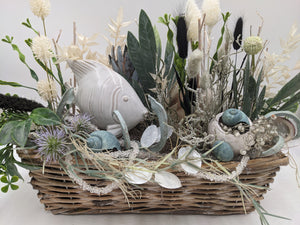 Nahaufnahme des Gestecks: weißer Fisch, Muscheln, Perlenzweige und violette Distel über dem Korbrand