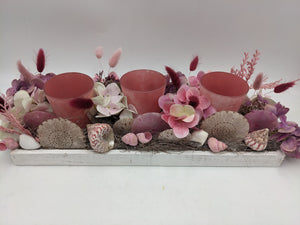 Tischgesteck in Rosa und Lila von vorn: drei rosafarbene Teelichtgläser zwischen Hortensienblüten, Schnecken und Muscheln
