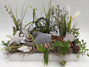 Maritimes Sommergesteck im weißen Holztablett mit grauem Fisch, Seestern, Muscheln, Gräsern und weißen Blüten