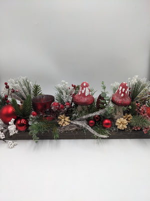 Langes Weihnachtsgesteck im dunklen Holztablett mit zwei roten Pilzen, Teelichtglas, roten Kugeln und beschneiten Zweigen