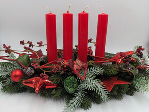 Langes Adventsgesteck mit vier roten Stabkerzen, roten Kugeln, Glassternen und Zapfen auf Tannenbeet
