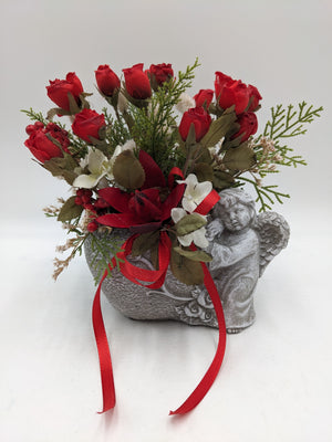 Graues Engelgefäß mit roten Rosen, weißen Blüten, Gräsern und roter Satinschleife von vorn