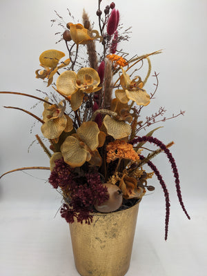 Hohes Herbstgesteck mit ockerfarbenen Orchideen, orangefarbener Schafgarbe und dunkelroten Gräsern im Goldtopf
