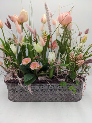 Frühlingsgesteck mit rosa und cremefarbenen Tulpen, Gräsern und Bellis in länglichem Metallkasten