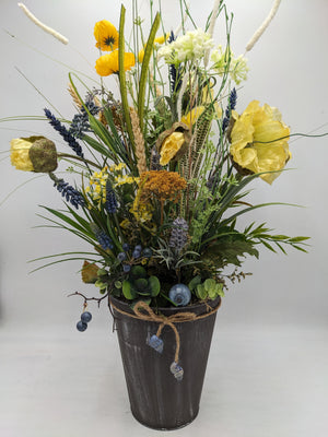 Hohes Frühlingsgesteck mit gelbem Mohn, blauen Hyazinthen und Gräsern in dunklem Topf mit Juteband