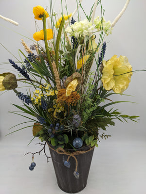 Frühlingsgesteck von vorn: gelbe Mohnblüten, ockerfarbene Schafgarbe, blaue Beeren und Ähren über dem Topfrand