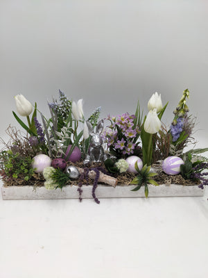 Längliches Ostergesteck in weißer Holzschale mit weißen Tulpen, lila Blüten, silbernem Hasen und Ostereiern