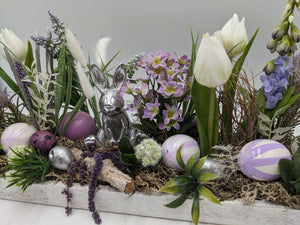 Nahaufnahme: silberner Hase zwischen lila-weiß gestreiften Ostereiern, Moos, Gräsern und weißen Tulpen