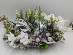 Frühlingsgesteck von vorn: silberne Hasenfiguren im Nest aus Wurzeln, weiße Blüten, Gräser und blaugraue Eier