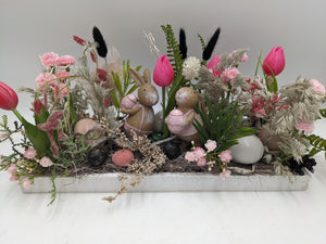 Längliches Ostergesteck im weißen Holzkasten mit zwei Hasenfiguren, pinken Tulpen, Gräsern und bunten Eiern