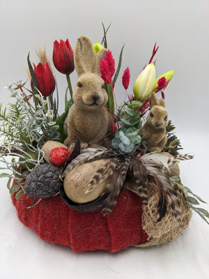 Ostergesteck mit zwei beigen Hasenfiguren, roten Tulpen, weißen Lilienknospen und Holzeiern auf rotem Filz