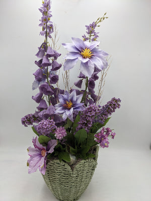 Hohes Sommergesteck in Lila mit Clematis, Glockenblüten und Flieder in geflochtenem graugrünem Korb