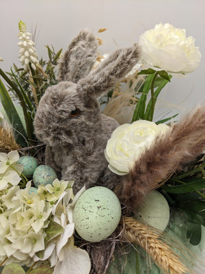 Nahaufnahme des grauen Hasen zwischen weißen Ranunkeln, Hortensienblüten, Ähren und mintgrünen gesprenkelten Eiern