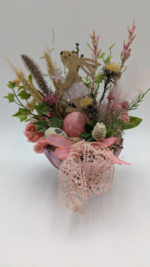 Ostergesteck mit Holzhase, rosa Ei, Gräsern, Federn und Spitzenband in rosa Glasschale, Nahaufnahme