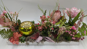 Frühlingsgesteck von vorn: rosa Tulpe, Protea, Gräser, Moos und grüne Libelle um ein goldgrünes Teelichtglas