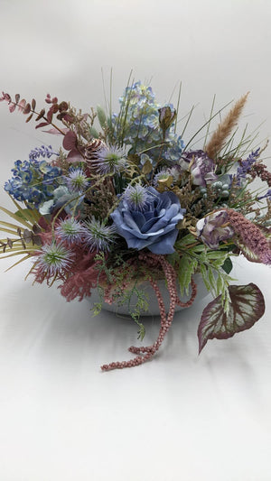 Herbstgesteck mit blauer Rose, blauen Hortensien, Disteln und Gräsern in heller Schale, seitlich fotografiert