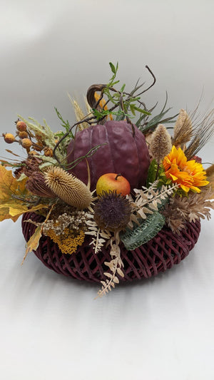 Herbstgesteck mit auberginefarbenem Kürbis, gelber Sonnenblume, Apfel und Hagebutten auf weinrotem Korbfuß