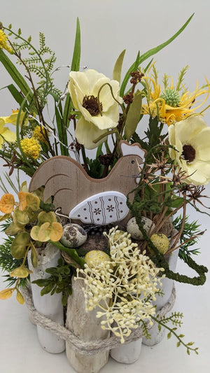 Nahaufnahme des Ostergestecks: Holzhuhn, gesprenkelte Eier, Moos und cremefarbene Beerendolden zwischen gelben Blüten