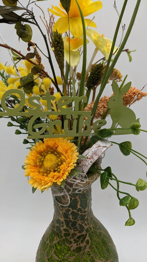 Nahaufnahme des Gestecks: gelbe Gerbera, Narzissen, Ähren, grüner Hase und Schriftzug Oster Grüße über der Vase