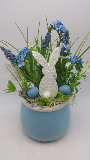 Ostergesteck mit weißem Glitzerhasen, blauen Eiern, Traubenhyazinthen und Gräsern in hellblauem Keramiktopf