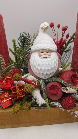 Nahaufnahme der Weihnachtsmannfigur mit weißer Glitzermütze zwischen roten Beeren, Geschenkpäckchen, Zapfen und Mohnkapseln