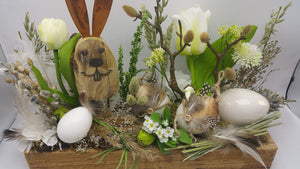 Nahaufnahme des Ostergestecks: Holzhase mit Rostohren, weiße Tulpen, Eier mit Federn und grünes Ei im Moos