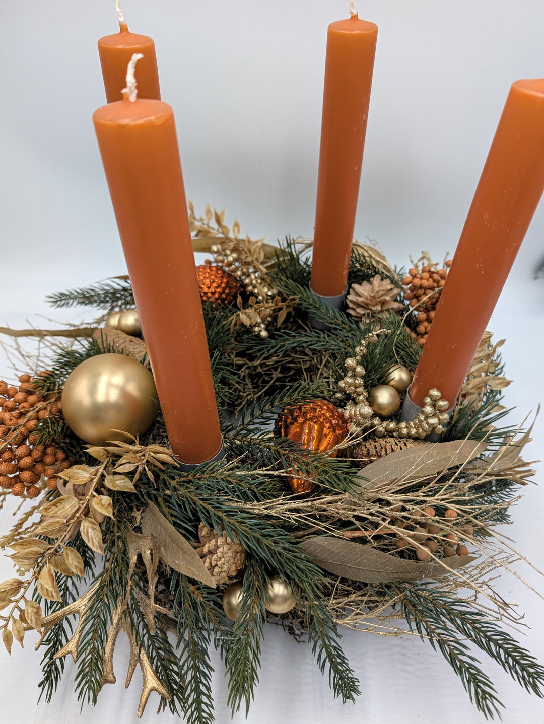 Adventskranz von schräg oben: orange Kerzen, große goldene Kugel, orange Glaskugel, Zapfen und getrocknete Gräser