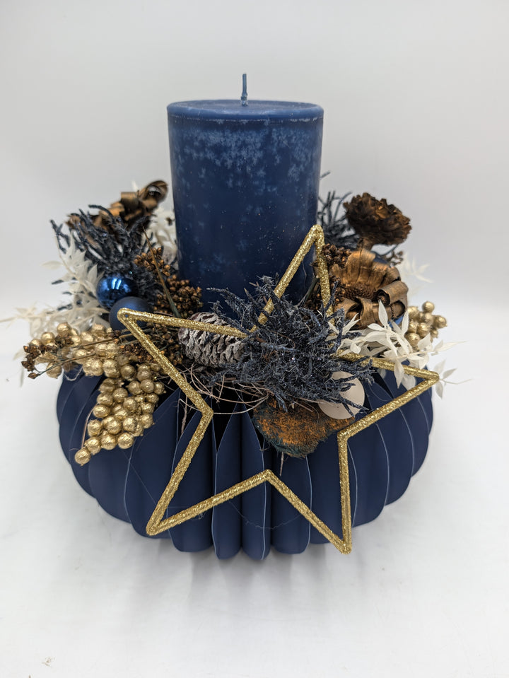 Weihnachtsgesteck mit blauer Stumpenkerze, goldenem Stern, Zapfen und goldenen Beeren auf dunkelblauem Gefäß