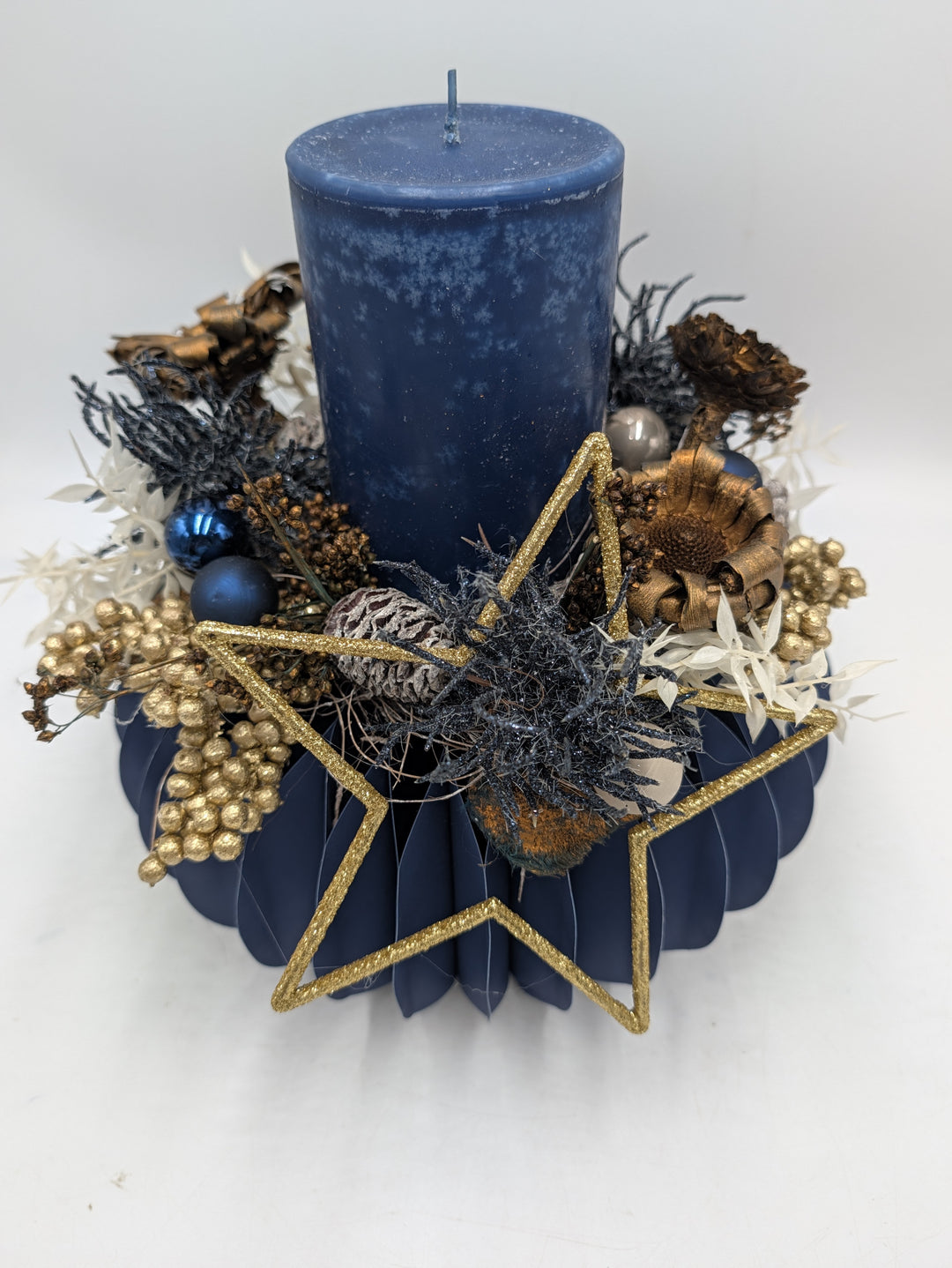 Gesteck von vorn: blaue Kerze, goldener Glitzerstern, kupferfarbene Blütenköpfe, blaue Kugeln und weiße Gräser