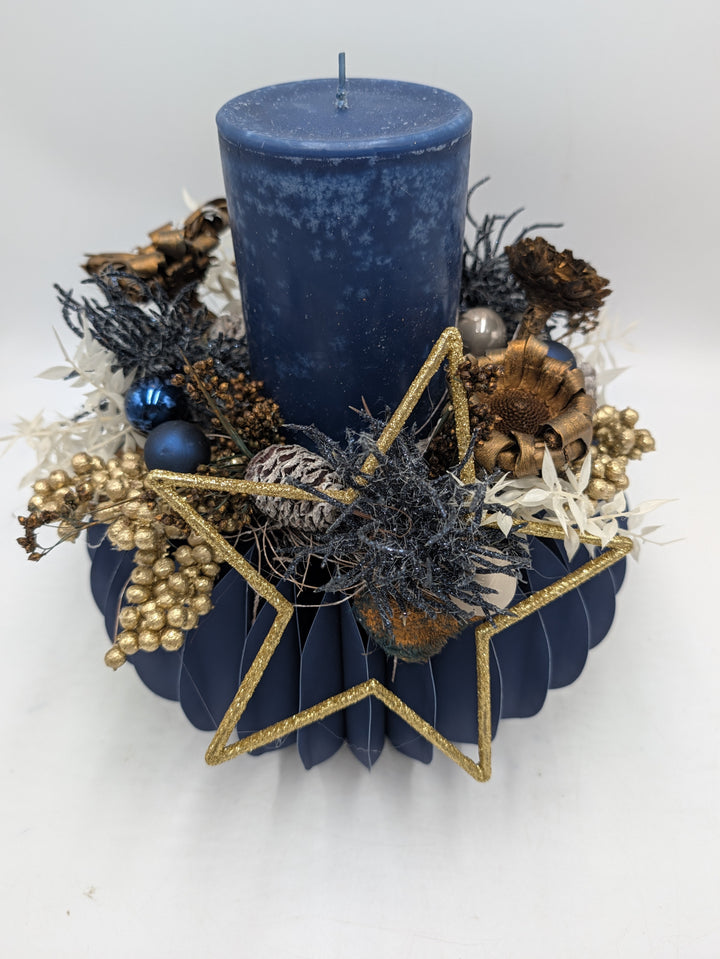 Gesteck von vorn: blaue Kerze, goldener Glitzerstern, kupferfarbene Blütenköpfe, blaue Kugeln und weiße Gräser