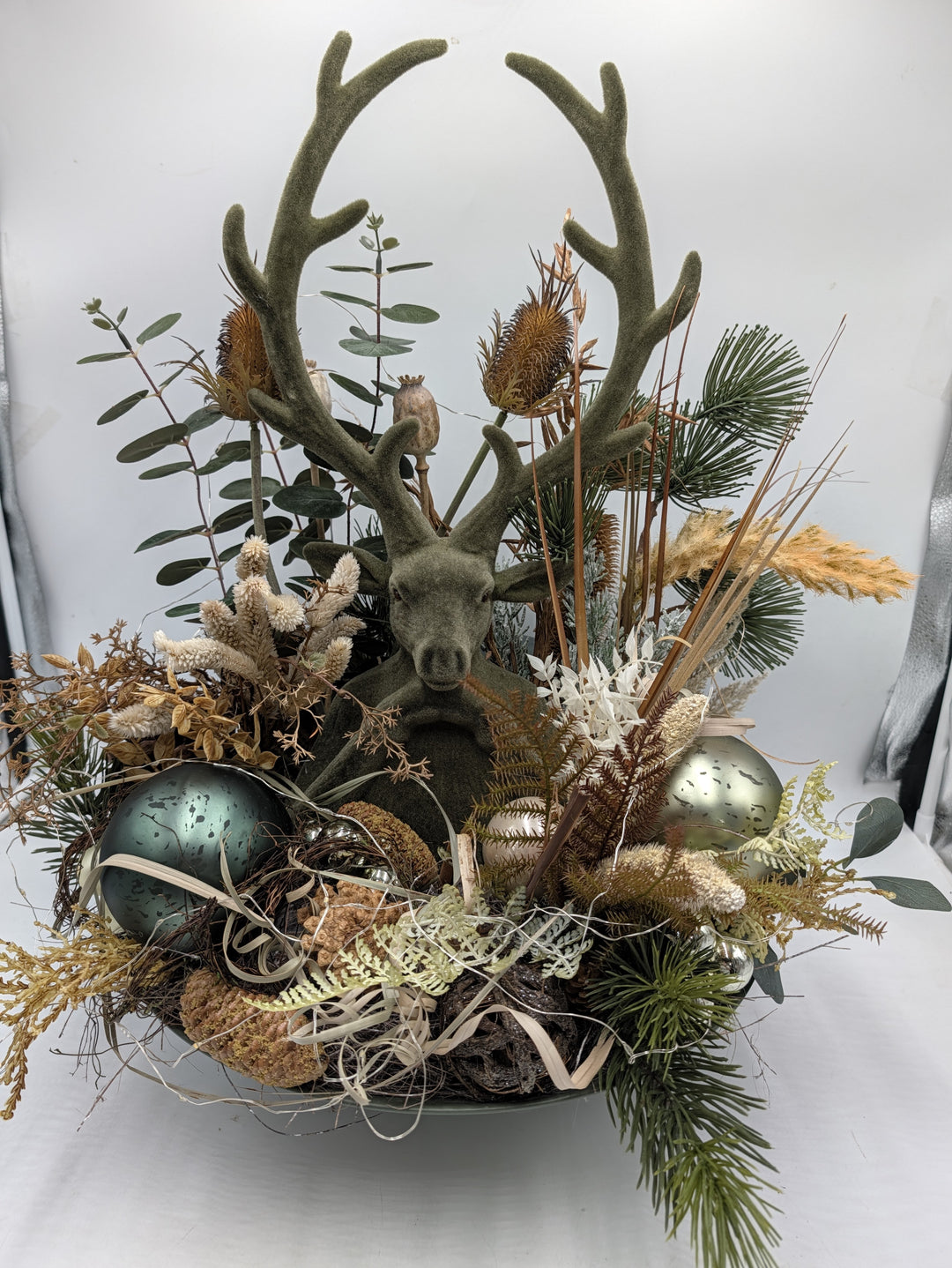 Wintergesteck mit grünem Hirsch, Karden, Eukalyptus und salbeigrünen Glaskugeln in flacher Schale