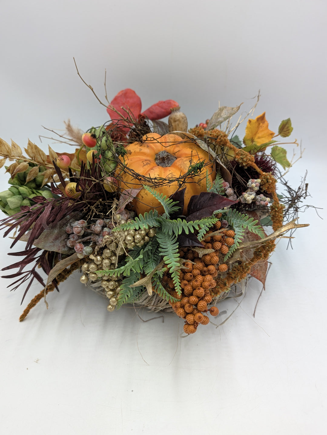 Flaches Herbstgesteck mit orangem Kürbis, grünen Hopfendolden, Farn, Beeren und Gräsern auf Zweigkranz