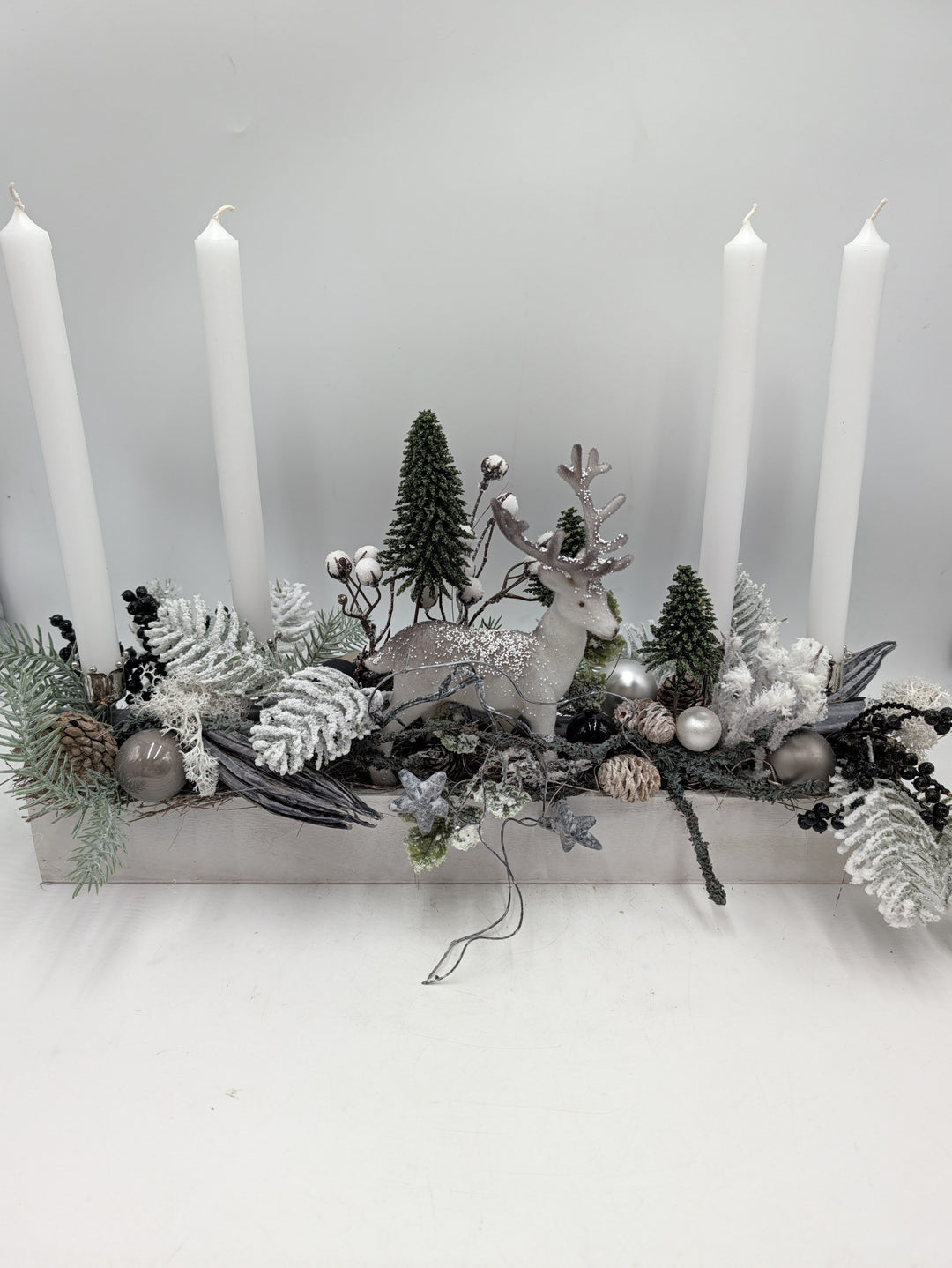Längliches Adventsgesteck in Weiß und Grau mit Hirsch, Tannenbäumchen und vier weißen Stabkerzen im Holzkasten