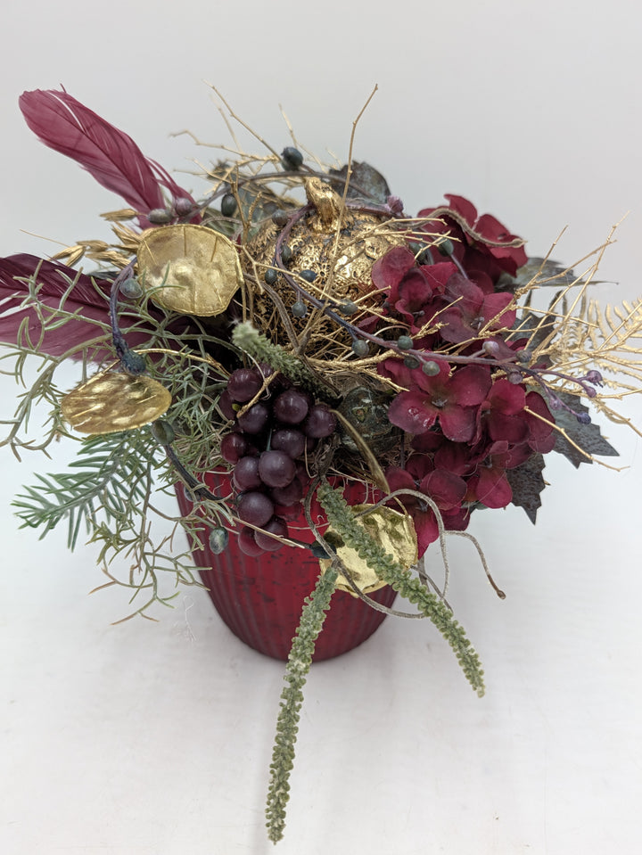 Herbstgesteck mit goldenem Kürbis, dunkelroten Hortensien, Trauben und bordeauxroten Federn im roten Topf
