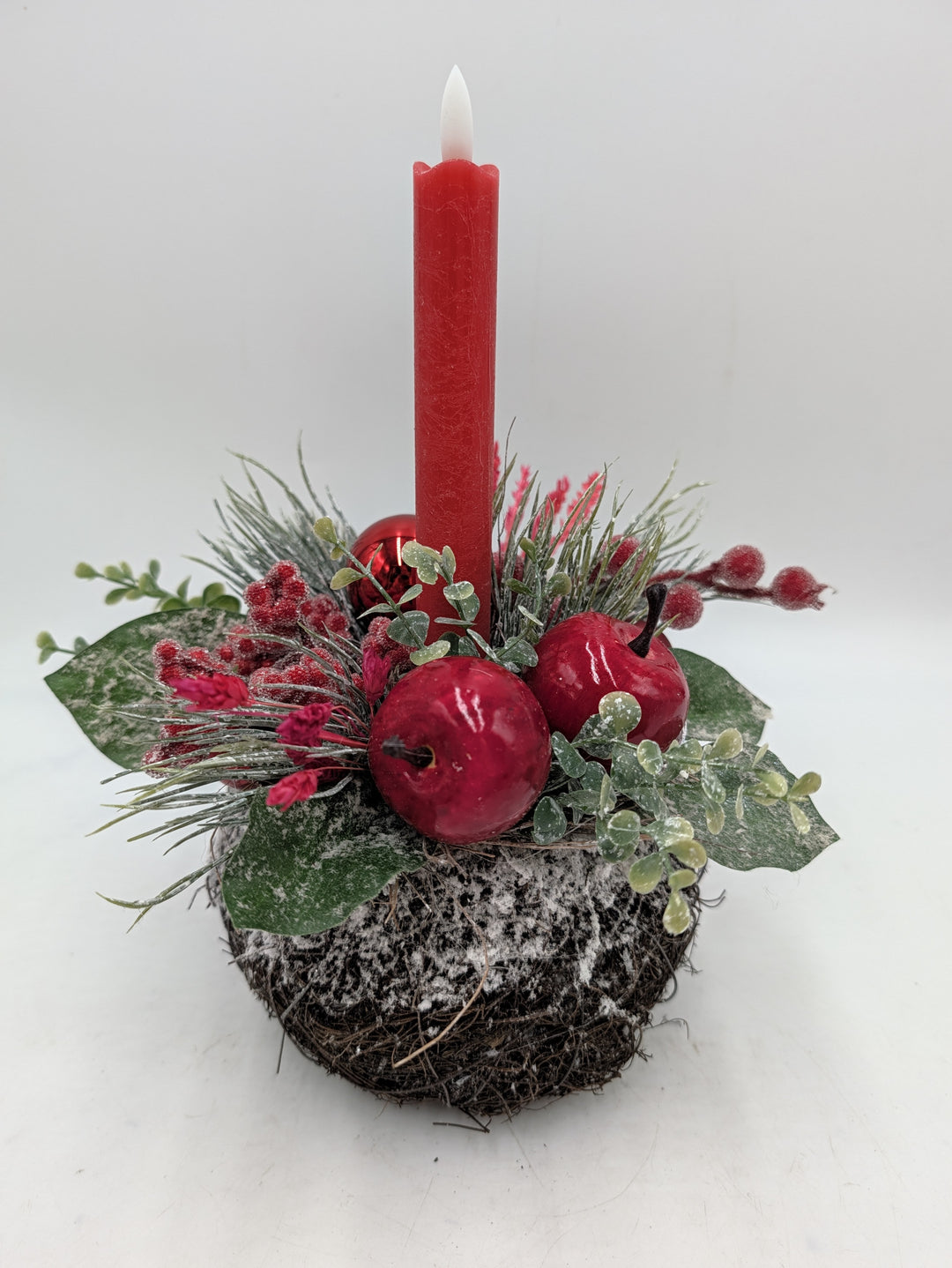 Weihnachtsgesteck mit roter LED-Stabkerze, zwei roten Äpfeln, Beeren und beschneiten Zweigen auf Reisigsockel