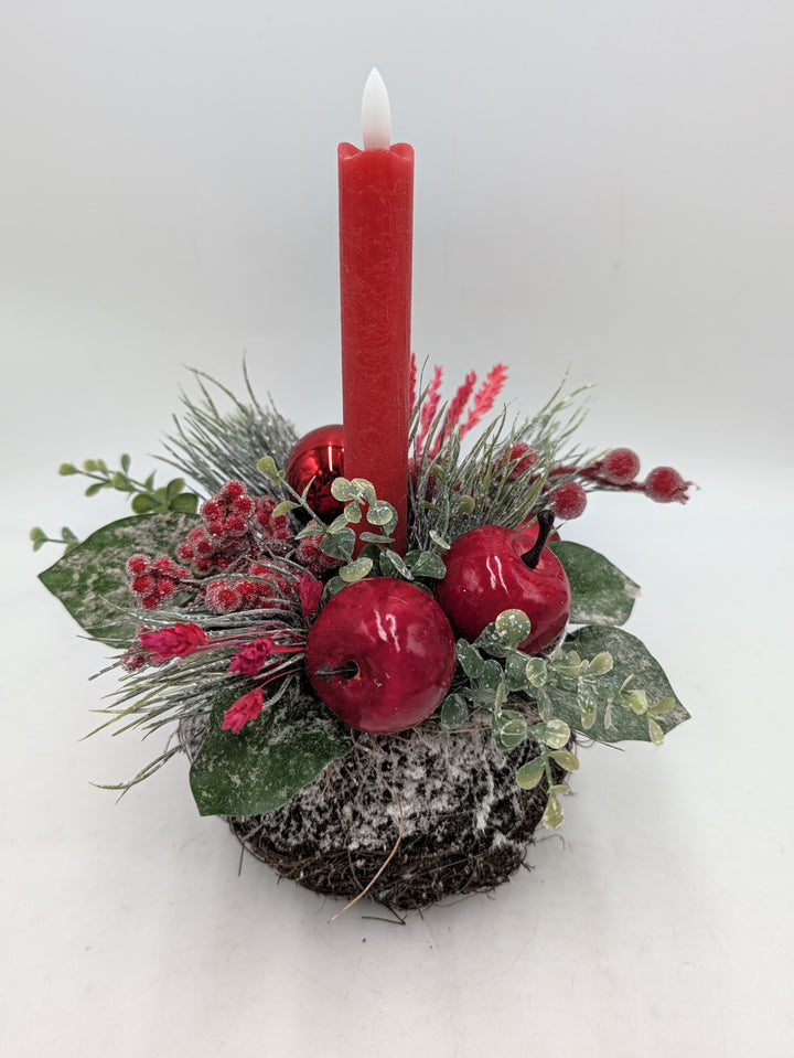 Weihnachtsgesteck von vorn: rote Kerze, Äpfel, rote Kugel, pinke Blütenähren und grüne Blätter mit Schneeeffekt